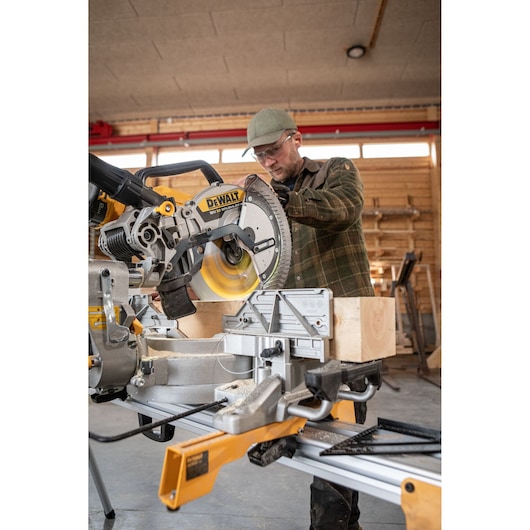 A DEWALT miter saw (model DCS782N) in use, cutting a wooden beam in a workshop environment. The saw is mounted on a stand and is operated by a person whose face is blurred for privacy.