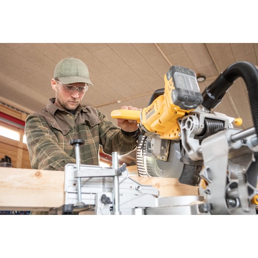 A person operating a DEWALT DCS782N sliding compound miter saw while cutting a piece of wood in a workshop. The saw is yellow and silver, and the person is wearing a plaid shirt and a cap. The face is blurred for privacy.