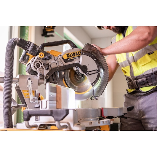 A DEWALT miter saw being used by a worker wearing safety gear to cut a piece of wood in an indoor construction setting.