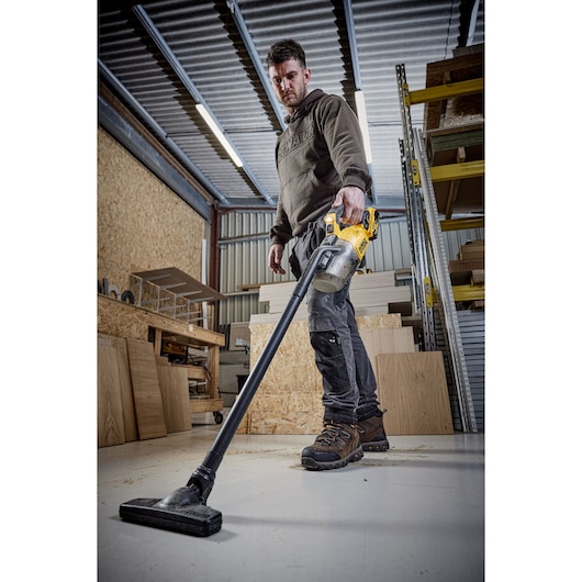 A person using a DEWALT cordless vacuum cleaner (model DCV501LN) to clean the floor in a workshop with wooden panels and shelving units.