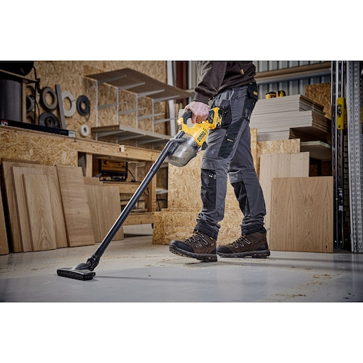 A person using a yellow DEWALT cordless vacuum cleaner to clean a workshop floor, surrounded by wooden boards and tools.