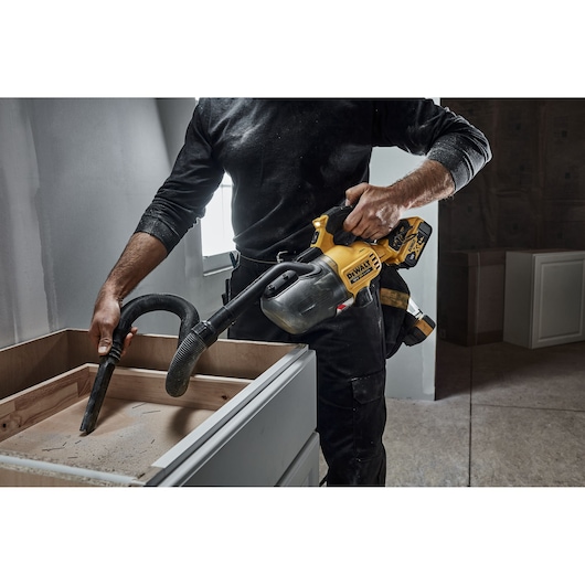 A person using a DEWALT cordless handheld vacuum cleaner (model DCV501LN) to clean sawdust from the surface of a wooden cabinet in a workshop or renovation setting.