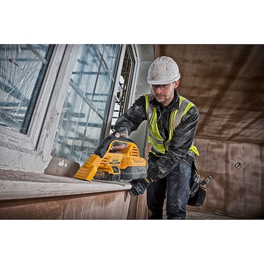 A construction worker wearing a safety helmet and high-visibility vest uses a yellow DEWALT DCV517N portable vacuum to clean dust from a window sill inside a building under construction.
