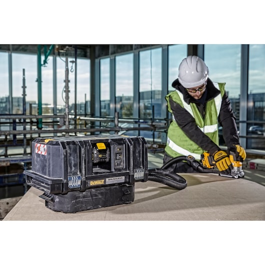 A DEWALT brushless dust extractor (model DCV586MT2) is placed on a work surface in a construction environment. In the background, a person wearing a safety helmet and high-visibility vest is using a power tool connected to the extractor.