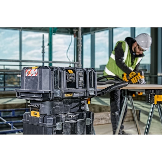 A DEWALT DCV586MT2 vacuum cleaner is pictured on a construction site, with a worker in safety gear sanding a wooden board in the background. The vacuum is connected via a hose to the working area.