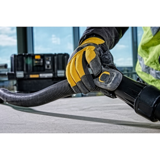 Close-up of a gloved hand connecting the hose to a DEWALT vacuum cleaner, with a DEWALT storage case visible in the background. The person is wearing a high-visibility jacket.