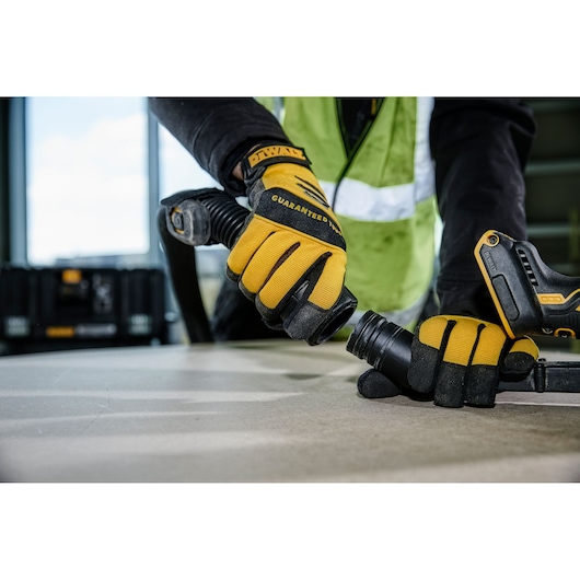Close-up of a person wearing yellow and black DEWALT branded gloves connecting a hose to a power tool on a work surface. A DEWALT toolkit is visible in the background.