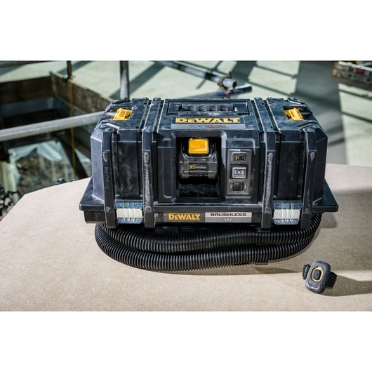 A DEWALT brushless 54V portable dust extractor with a remote control attachment, featuring a black and yellow design. The unit is placed on a work surface in an industrial setting.