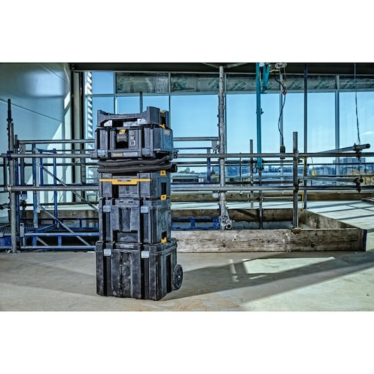 A stack of black and yellow DEWALT tool storage boxes with wheels, arranged on a construction site indoors. The boxes are part of an organized rolling system, standing in front of metal scaffolding and large glass windows.
