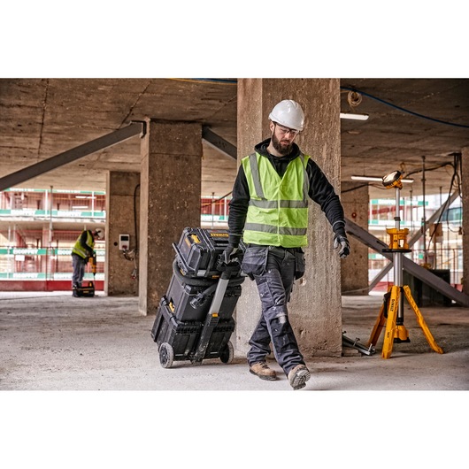 A construction worker wearing a safety helmet and high-visibility vest pulls a DEWALT wheeled toolbox through a building