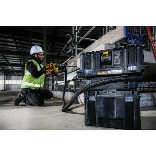 A DEWALT DCV586MT2 brushless vacuum with hose is placed on the floor at a construction site. In the background, a construction worker wearing a high visibility vest and helmet is using a yellow DEWALT power tool connected to the vacuum. Industrial scaffolding and concrete structures are visible.