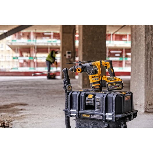 A DEWALT cordless rotary hammer drill with attached dust extraction system is placed on top of a DEWALT storage box on a construction site. In the background, a person in a safety vest is working.