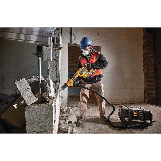 A construction worker wearing safety gear uses a DEWALT cordless dust extractor and rotary hammer to demolish a concrete block wall. The connected dust extractor with hose is placed on the floor, and debris is scattered around the work area.