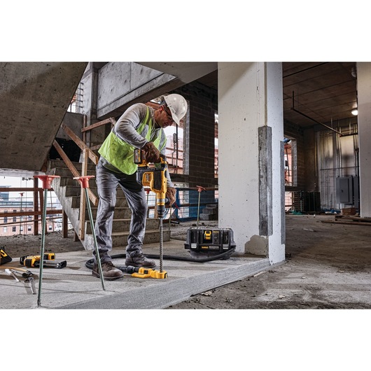 A worker in a construction site wearing a safety vest and helmet is using a DEWALT cordless power tool to drill into a concrete surface. The DEWALT DCV586MT2 dust extractor is visible on the floor beside the worker.