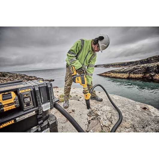 A construction worker wearing safety gear drills into a rocky surface by the water using a DEWALT power tool connected to a mobile DEWALT dust extractor. The scene is outdoors by a rocky shoreline under cloudy skies.