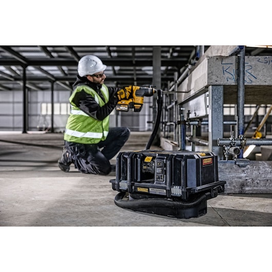 A DEWALT DCV586MT2 brushless portable vacuum is placed on the floor at a construction site, with a worker in safety gear using a power tool in the background.