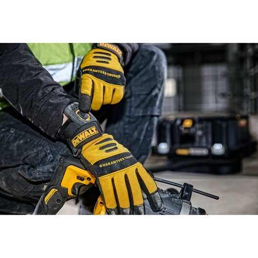 Close-up of a person wearing yellow and black DEWALT gloves, adjusting the wrist strap on one glove while holding a yellow DEWALT power tool. A DEWALT equipment case is visible in the background in an industrial setting.