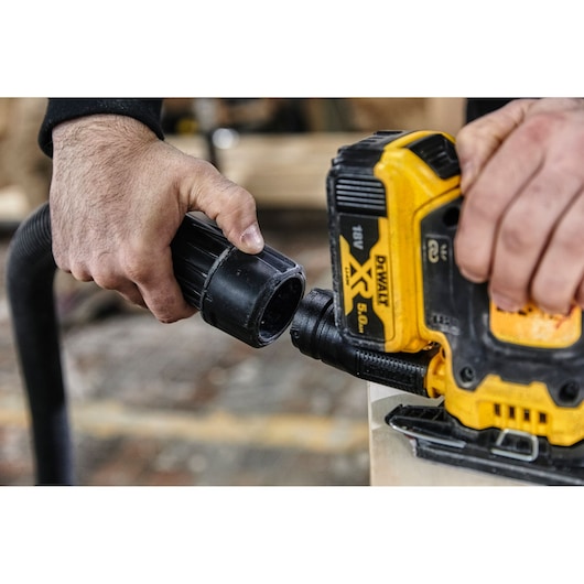Close-up of two hands connecting a vacuum hose to a yellow DEWALT power tool, with visible branding and model information.