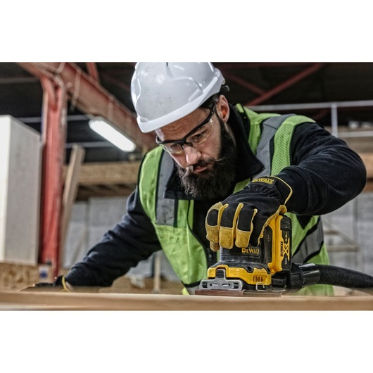 A person wearing a safety vest, gloves, and a white hard hat is using a yellow DEWALT DCW200N cordless sander on a wooden surface in an indoor workshop or construction site.