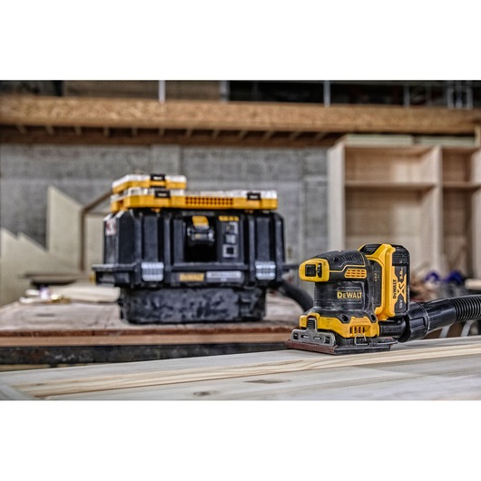 A DEWALT cordless electric sander is placed on a wooden surface in a workshop. In the background, there is a black and yellow DEWALT tool storage box. The setting appears to be a woodworking or construction area.