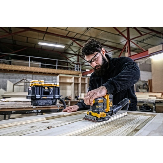 A person is using a DEWALT DCW200N cordless sander on a wooden surface in a woodworking workshop. The person's face is blurred for privacy. Various tools and wooden materials are visible in the background.