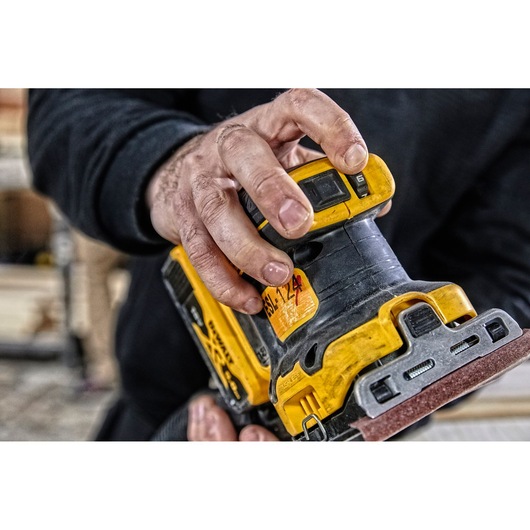 Close-up of a person's hand operating a DEWALT DCW200N cordless sander in a workshop environment.