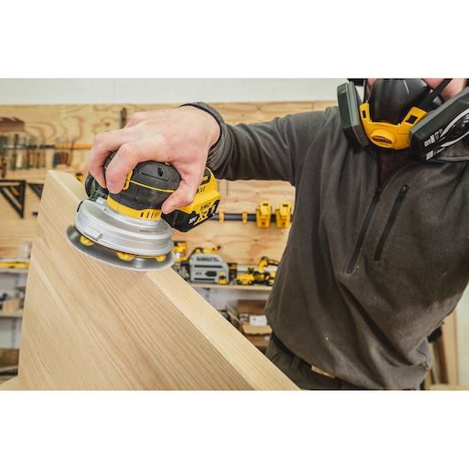 Person uses a DEWALT cordless sander on a wooden surface in a workshop, wearing a protective mask.