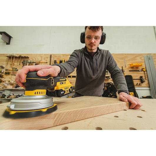 Person sanding wood using a DEWALT DCW230N-XJ cordless sander in a workshop.