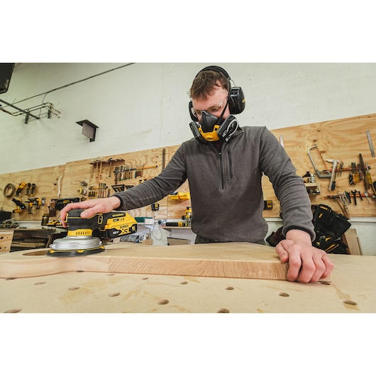Person sanding wood with a DEWALT sander, wearing safety gear in a workshop with tools on the wall.