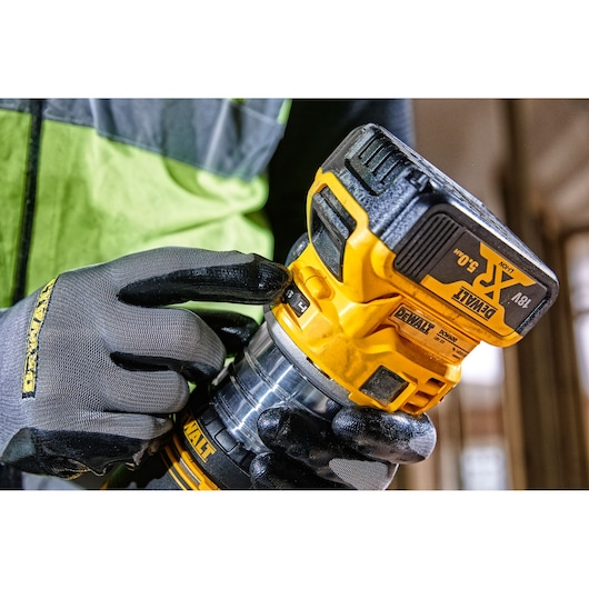 Worker uses DEWALT DCW604 cordless router, wearing gloves and adjusting the tool for woodworking.