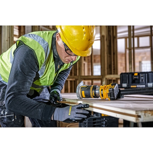 Worker uses DEWALT DCW604 router tool on wood in a construction site, wearing gloves, helmet, and safety vest.