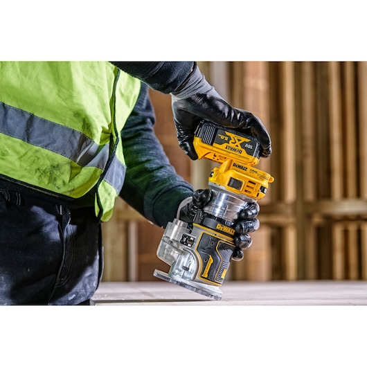 Worker uses a DEWALT DCW604 cordless router tool on a wooden surface at a construction site.