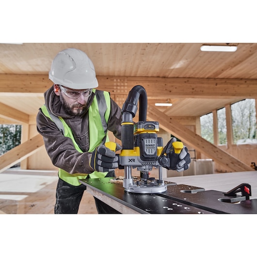 A construction worker wearing safety gear operates a DEWALT cordless router (model DCW620H2) on a wooden surface in a modern wood-framed building.