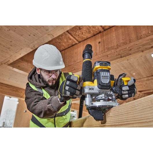 A person wearing safety gear operates a DEWALT XR cordless router on a wooden beam inside a construction site. The DEWALT tool is prominently visible, and wood shavings are flying as the router is used.