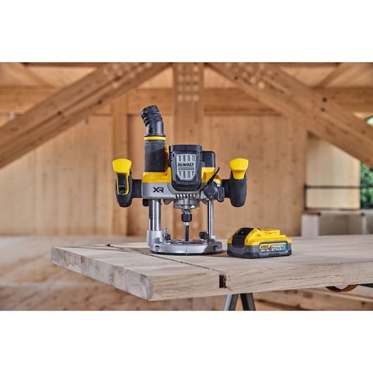 DEWALT cordless router with yellow and black design placed on a wooden table in a woodworking workshop. A DEWALT Power Stack battery is positioned next to the router.