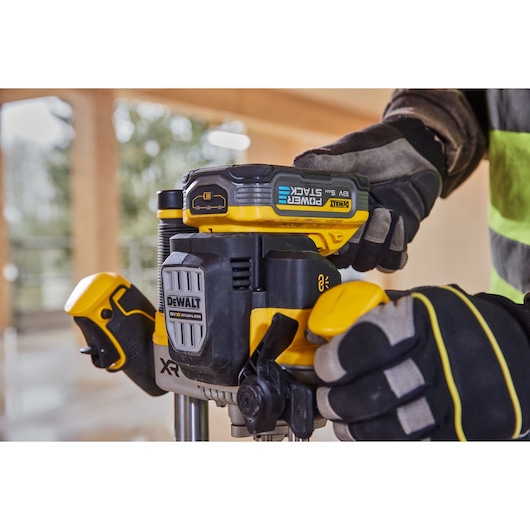 Close-up of a person using a DEWALT DCW620H2 cordless router with a POWERSTACK lithium battery, wearing protective gloves, in a construction setting.
