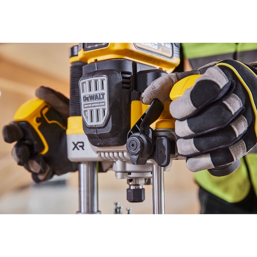 Close-up view of a person wearing work gloves operating a DEWALT cordless power tool, specifically a router, indoors.