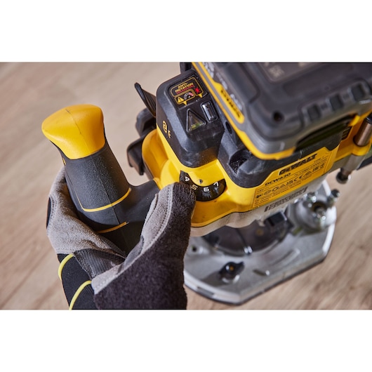 Close-up of a gloved hand adjusting the speed dial on a DEWALT DCW620H2 router, with visible yellow and black handle and control panel, placed on a wooden surface.