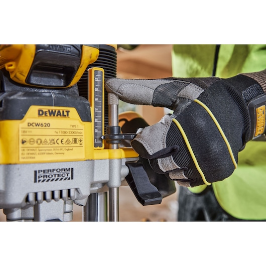 Close-up of a person wearing protective gloves adjusting the depth gauge on a DEWALT DCW620 power tool. The yellow and black DEWALT branding is visible near the gauge and controls.