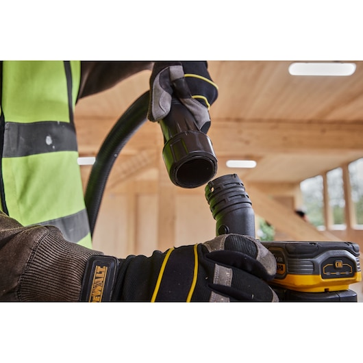 Close-up of a person wearing gloves connecting two black hoses to a DEWALT portable device in a wooden indoor workspace.