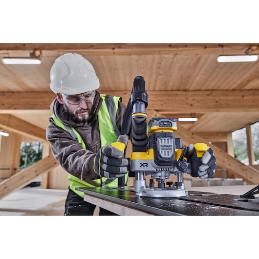A person wearing a safety helmet, gloves, and high-visibility vest is operating a DEWALT cordless router (DCW620NT) on a wooden surface inside a construction site with wooden beams.