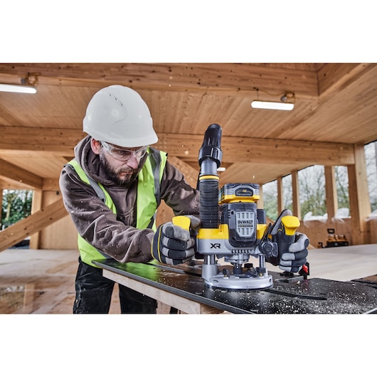 A person wearing a safety helmet and high-visibility vest is using a DEWALT DCW620NT cordless router on a wooden surface inside a timber building. The router is featured prominently with dust extraction attached, and the environment is well lit with wooden beams and windows.