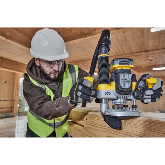 A person wearing a safety helmet and high-visibility vest operates a DEWALT XR cordless router (model DCW620NT) on a wooden plank in a construction environment.