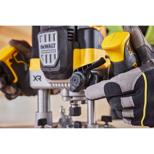 Close-up of a person wearing gloves adjusting a DEWALT XR brushless router tool. The focus is on the yellow and black tool's handle and adjustment lever.