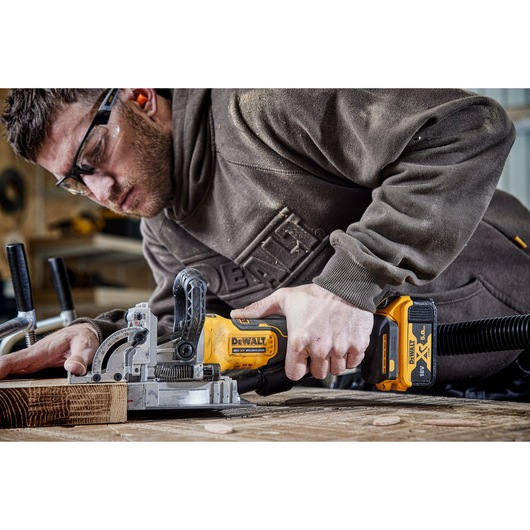 A person is using a DEWALT cordless biscuit joiner tool to work on a piece of wood in a workshop. The tool is yellow and black with a DEWALT XR 18V battery attached.