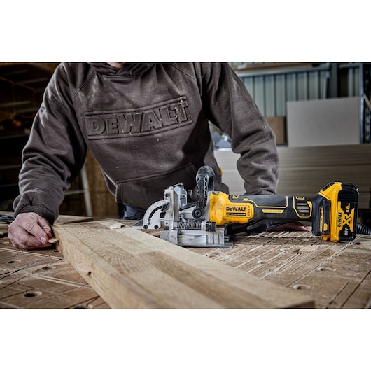 A person using a DEWALT cordless joiner tool to work on a wooden board in a workshop setting.