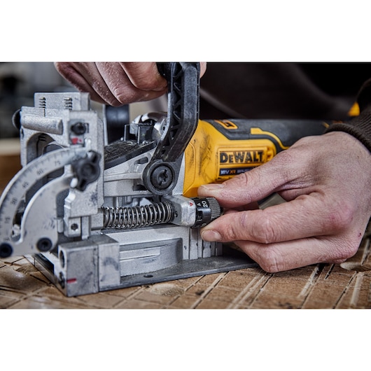 Close-up of a person adjusting a DEWALT power tool, specifically a biscuit joiner, with visible hands working on the adjustment knob and lever. The tool is placed on a wooden surface.