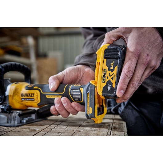 Close-up of a person inserting an 18V 5.0Ah DEWALT XR battery into a cordless DEWALT power tool, placed on a wooden surface.