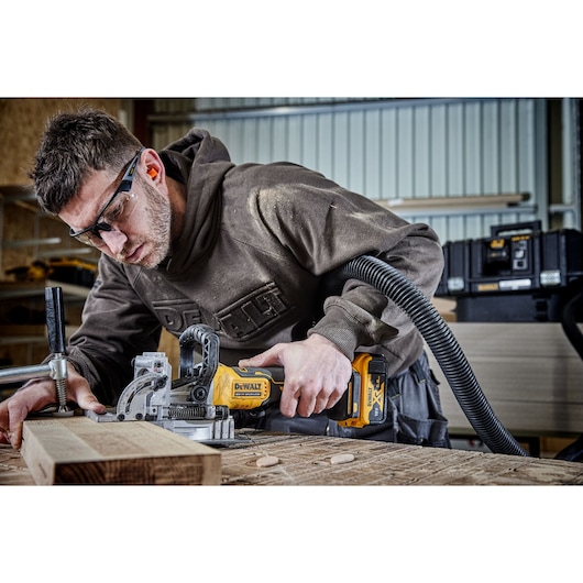 A person is using a DEWALT DCW682P2 cordless joiner tool on a wooden workpiece in a workshop, with wood dust visible and a vacuum hose attached for dust extraction.