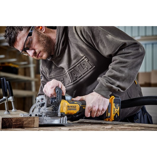 A person using a DEWALT cordless biscuit joiner (model DCW682P2) to work on a piece of wood in a workshop, with clamps securing the wood on the table. The person is wearing a DEWALT sweatshirt, and the joiner is attached to a dust extraction hose.